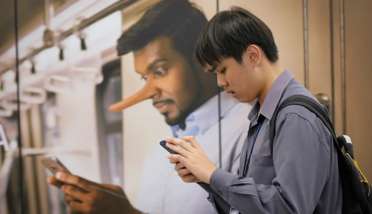 Penumpang kereta bermain handphone dengan latar belakang iklan larangan hoax di Stasiun Kereta Kuala Lumpur, Malaysia, Senin (2/4). Pengesahan UU Antihoax dikhawatirkan membungkam kelompok yang berseberangan dengan pemerintah. (AP Photo/Vincent Thian)