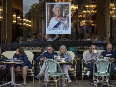 Orang-orang duduk di bar teras di sebelah potret Ratu Elizabeth II di pusat kota London pada 13 September 2022. Hotel, restoran, dan toko-toko penuh sesak saat penggemar kerajaan berdatangan ke jantung kota London untuk melihat jalan-jalan yang dipagari bendera, prosesi yang dipenuhi kemegahan, dan berani mengantre sejauh bermil-mil untuk kesempatan sekali seumur hidup mengucapkan selamat tinggal kepada Ratu Elizabeth II. (AP Photo/Emilio Morenatti, File)