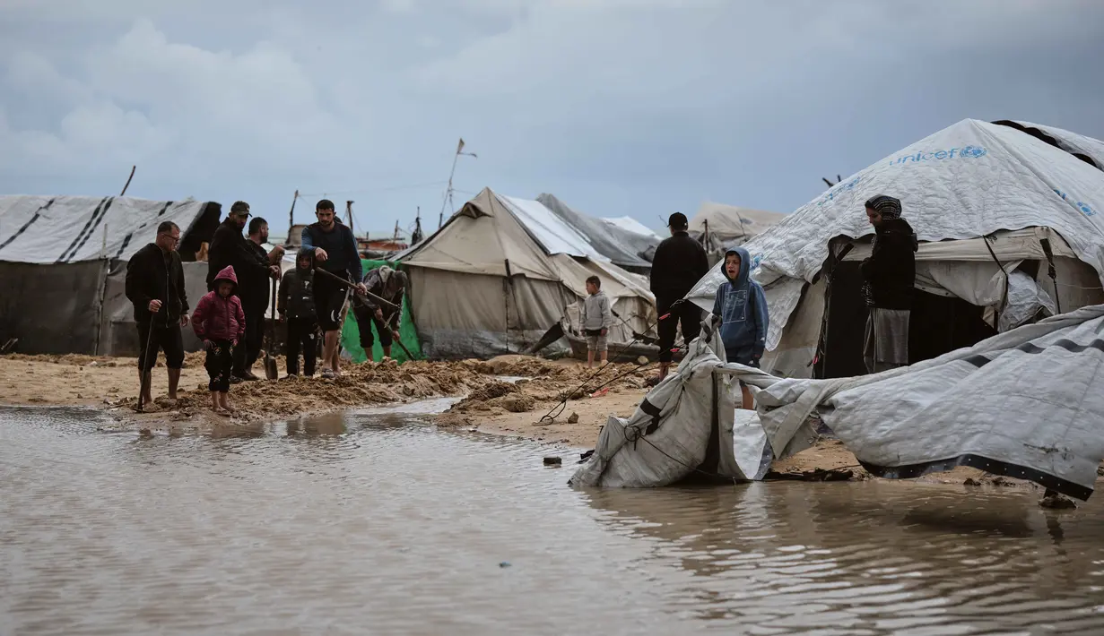 Hujan deras dan angin kencang merendam tenda-tenda para pengungsi, dan memperburuk krisis kemanusiaan yang sudah ada. Tampak dalam foto, warga Palestina berjalan melewati area yang tergenang banjir di kamp tenda sementara setelah hujan lebat di Kota Gaza, Kamis, 26 Maret 2026 (AP Photo/Jehad Alshrafi)