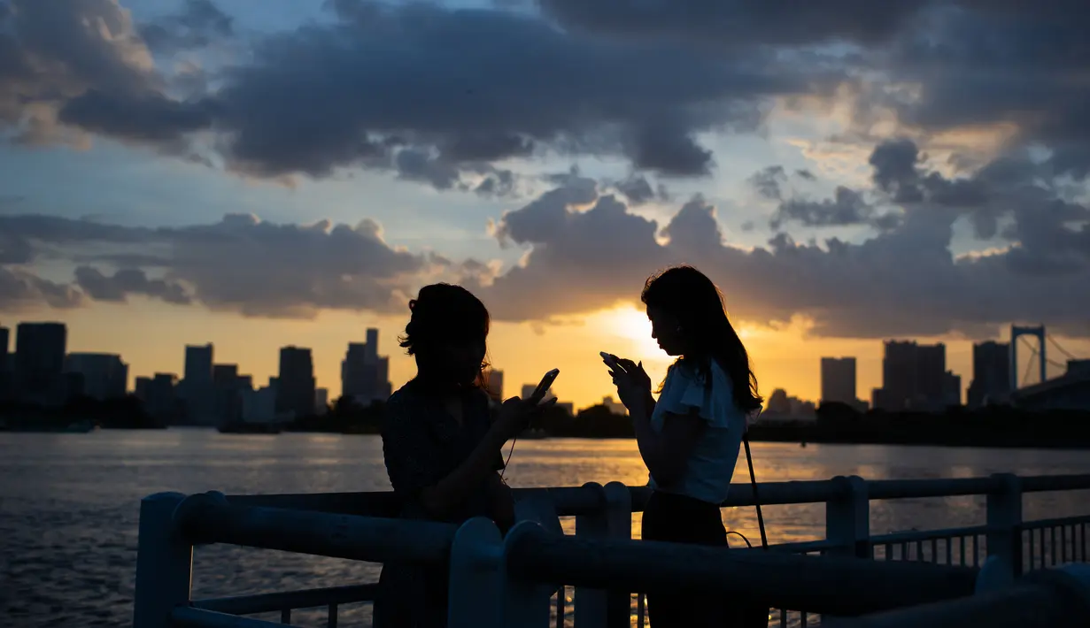 Dua wanita bermain ponsel mereka saat matahari terbenam di Odaiba, Tokyo, (29/7). Nama "Odaiba" berasal dari sebutan untuk enam pulau di Teluk Tokyo yang dibangun sebagai benteng oleh Keshogunan Tokugawa. (AFP Photo/Martin Bureau)
