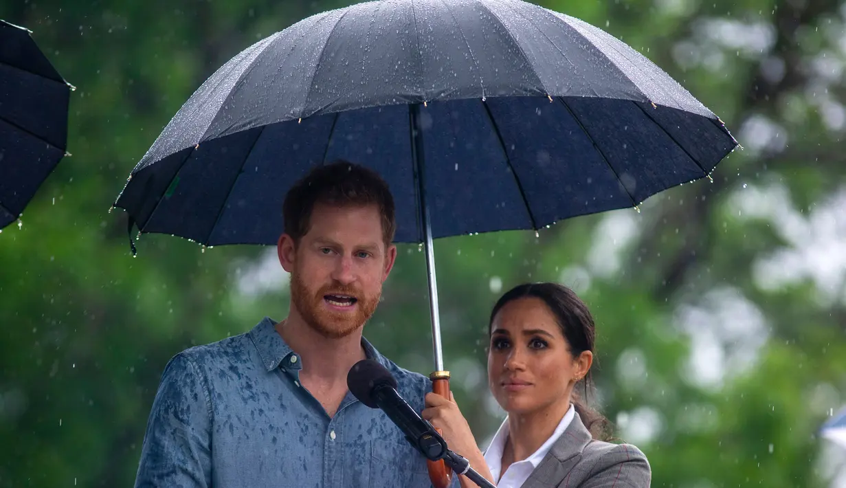 Meghan Markle memayungi Pangeran Harry saat memberi sambutan dalam kunjungan ke Victoria Park, Dubbo, Australia, Rabu (17/10). Ini adalah hari kedua Harry dan Markle dalam kunjungannya ke Australia. (Ian Vogler/Pool via AP)