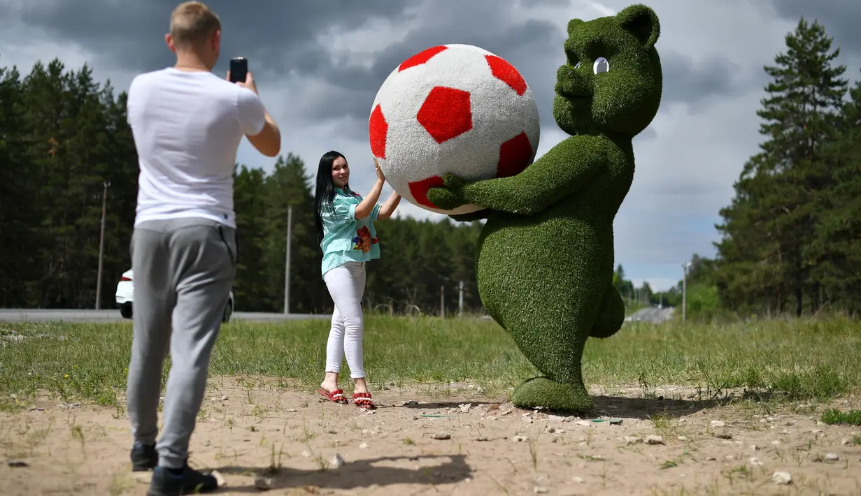Seorang wanita berpose di samping patung seekor anak beruang memegang bola yang terbuat dari dedaunan di dekat bandara Samara, Rusia (11/6). Rusia akan menjadi tuan rumah turnamen sepak bola Piala Dunia 2018. (AFP Photo/Fabrice Coffrini)