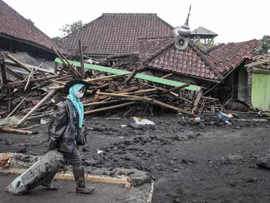 Seorang perempuan melintas di depan sebuah rumah yang hancur akibat aliran piroklastik Gunung Semeru di Desa Supiturang, Lumajang, Jawa Timur, pada Jumat 21 November 2025. Hari ketiga pasca-erupsi Gunung Semeru, atau Jumat (21/11/2025), Pemerintah Desa Supiturang, Kecamatan Pronojiwo, Kabupaten Lumajang, Jawa Timur, mencatat sedikitnya lebih dari 200 rumah dan fasilitas umum rusak parah. (Agus Harianto/AFP)