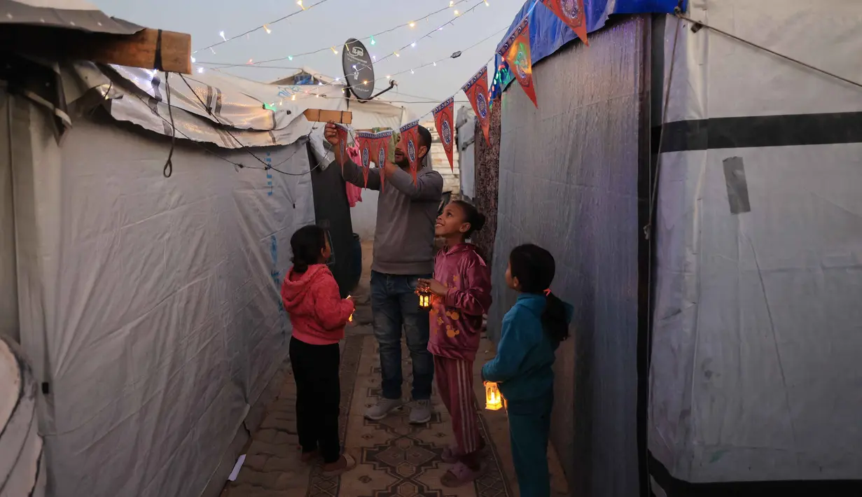 Momen tersebut menjadi ruang kecil untuk merasakan kebersamaan di tengah situasi yang belum sepenuhnya pulih. Tampak dalam foto, Para pengungsi muda Palestina memegang lampion Ramadan saat dekorasi dipasang di samping tenda mereka di kamp pengungsi Bureij di Jalur Gaza tengah pada 15 Februari 2026. (Eyad Baba/AFP)