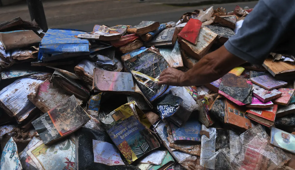 Seorang pria mengambil buku rusak yang ditinggalkan di jalan setelah hujan lebat membanjiri toko buku di Petropolis, Brasil, 19 Februari 2022. Sebanyak 136 jenazah telah diidentifikasi hingga saat ini, menurut pejabat pertahanan sipil. (MAURO PIMENTEL/AFP)