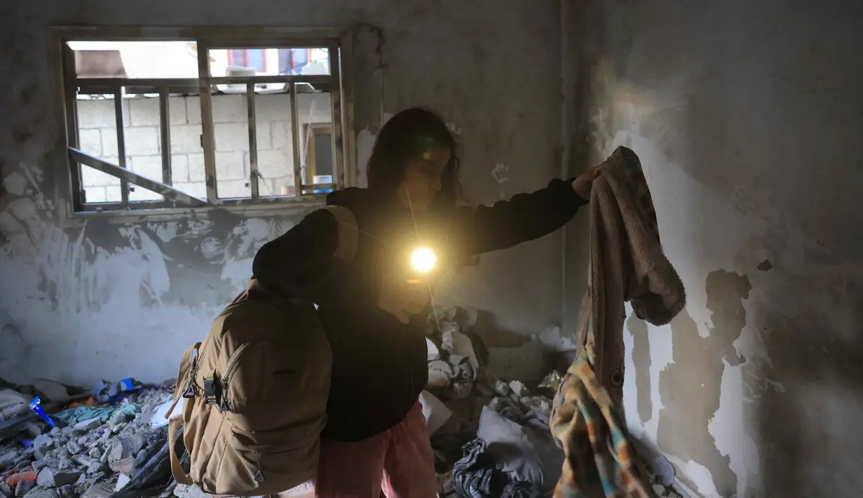 Sementara, jumlah korban luka meningkat drastis hingga melebihi 171.324 orang. Tampak dalam foto, seorang wanita Palestina menggunakan senter di ponselnya untuk mengambil pakaian dari bawah reruntuhan di sebuah ruangan, setelah serangan militer Israel terhadap rumah keluarga al-Houli, di mana empat orang dilaporkan tewas, di sebelah barat Deir al-Balah, di Jalur Gaza tengah pada Jumat 16 Januari 2026. (BASHAR TALEB/AFP)