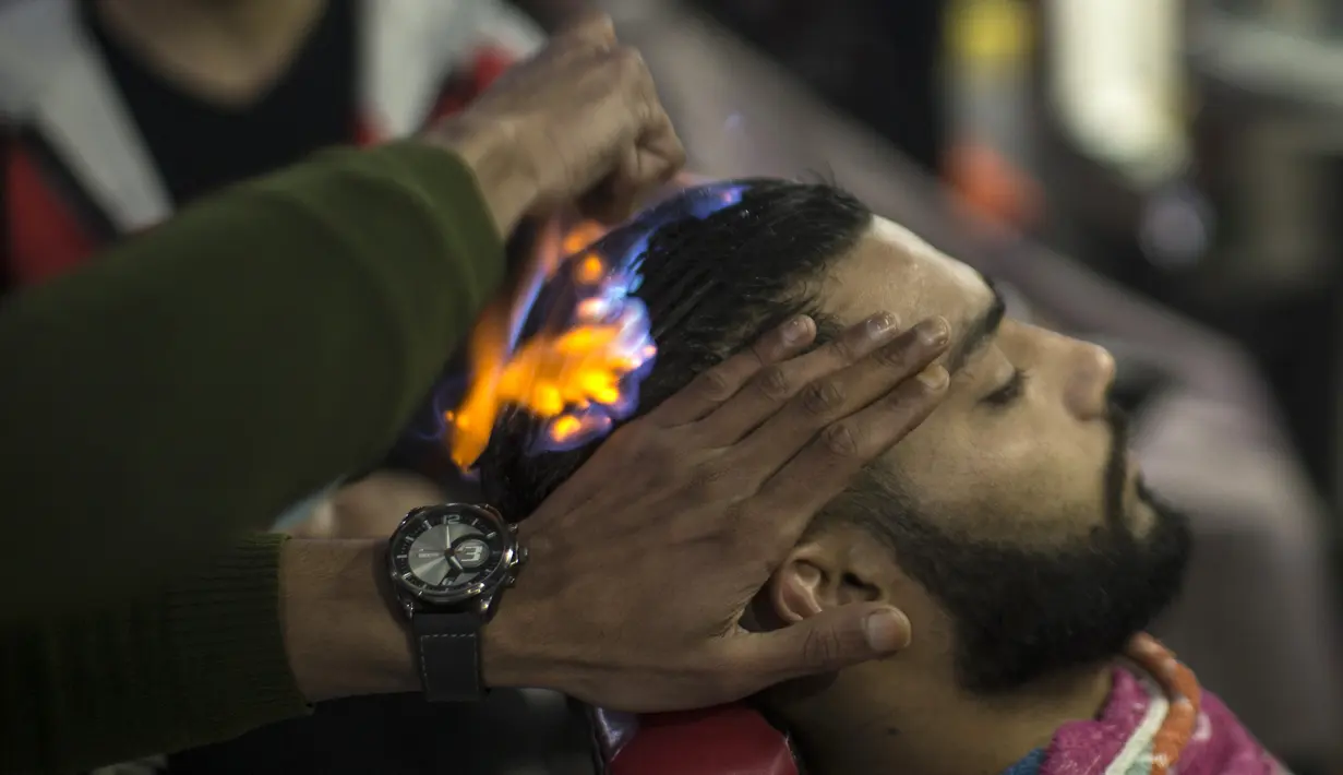 Ramadan Edwan, seorang tukang cukur asal Palestina, dengan cekatan memotong rambut pelanggannya yang terbakar oleh api di sebuah barbershop di kamp pengungsian Rafah, di selatan Jalur Gaza, Rabu (1/2). (MAHMUD HAMS/AFP)