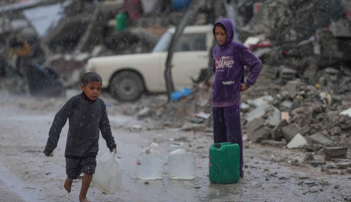 Rata-rata anak-anak di Gaza hanya mendapatkan 1,5 hingga 2 liter air per hari, jauh di bawah kebutuhan dasar manusia. Tampak dalam foto, seorang anak laki-laki tanpa sepatu berjalan di bawah hujan sambil membawa jerigen plastik berisi air di kawasan Sheikh Radwan, Kota Gaza, pada Jumat 14 November 2025. (AP Photo/Jehad Alshrafi)