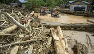 Tumpukan material kayu yang terbawa air saat banjir bandang tidak hanya menutup jalan, tetapi juga menimbun rumah dan halaman warga. Tampak dalam foto, warga desa mengarungi banjir di jalan yang tergenang akibat banjir bandang di Desa Tukka, Tapanuli Tengah, Sumatera Utara, pada 2 Desember 2025. (YT HARIONO/AFP)