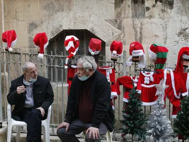 Orang-orang duduk di depan sejumlah kostum Sinterklas yang dipajang di dekat sebuah toko di Kota Tua Yerusalem (7/12/2020). (Xinhua/Muammar Awad)