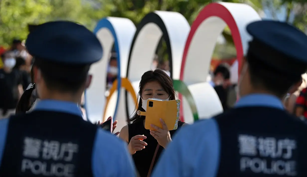 Polisi berjaga saat orang-orang berpose di depan cincin Olimpiade jelang upacara pembukaan Olimpiade Tokyo 2020 di luar Stadion Olimpiade, Tokyo, Jepang, Jumat (23/7/2021).  Upacara pembukaan Olimpiade Tokyo 2020 akan digelar pada 23 Juli 2021 malam. (Andrej ISAKOVIC/AFP)