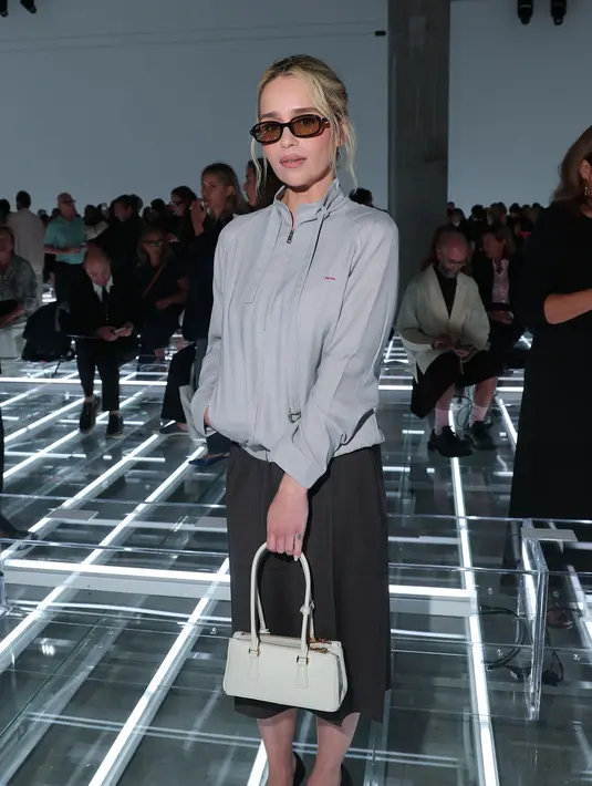 Emilia Clarke tampil dengan atasan zip abu-abu muda, dipadukan rok abu-abu gelap. Kacamata cokelat, tas Prada putih, dan heels slingback hitam lengkapi busananya [dok. Prada]
