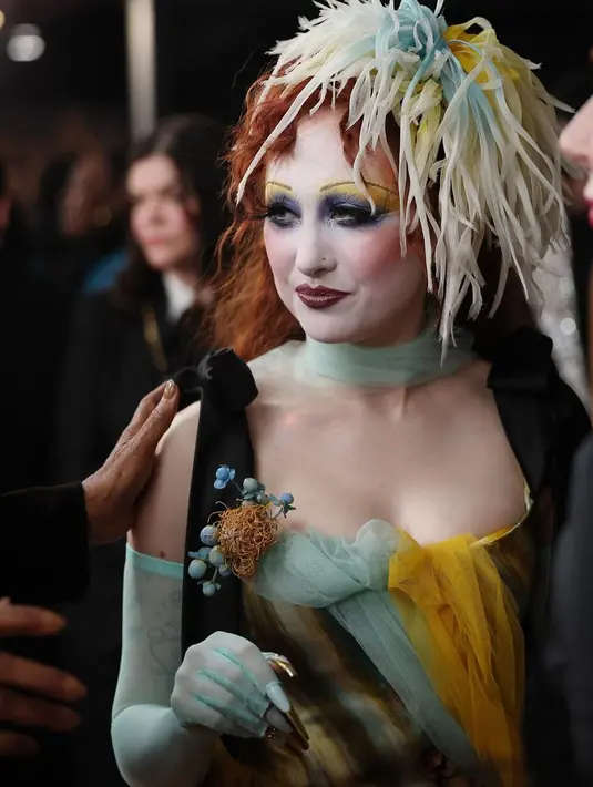 Gaun ini juga dihiasi dengan sarang ranting dan bunga biru di sepanjang garis leher dan pinggang. Untuk melengkapi penampilannya, ia mengenakan sepatu bot kulit lace-up rancangan John Fluevog, sarung tangan opera transparan tanpa jari, serta hiasan topi berbulu warna-warni yang serasi. [@genesiswbb].