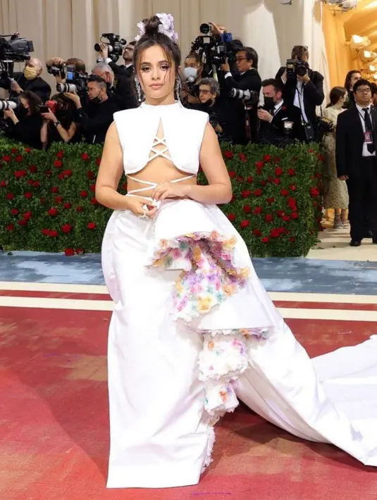 <p>Camila Cabello, mengenakan gaun putih dengan ruffles bunga yang tidak menarik di atasnya. Namun sayangnya, detail kereta panjang di pinggulnya membuat perutnya terlihat penuh. (Instagram/metgalaofficial).</p>
