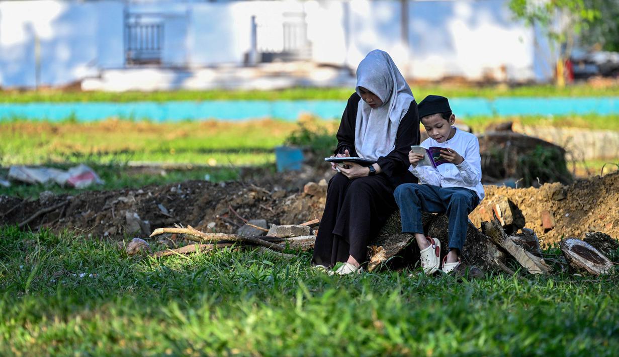Bagi masyarakat Aceh, momen peringatan tragedi tsunami kali ini bukan hanya mengenang kehilangan masa lalu, tetapi juga tentang menghadapi tantangan masa kini. Tampak dalam foto, seorang wanita dan anaknya membaca Al-Quran sambil berdoa untuk kerabat yang meninggal dunia dalam peristiwa gempa dan tsunami Aceh pada 2004 lalu di sebuah kuburan massal di Siron, pada 26 Desember 2025. (CHAIDEER MAHYUDDIN/AFP)