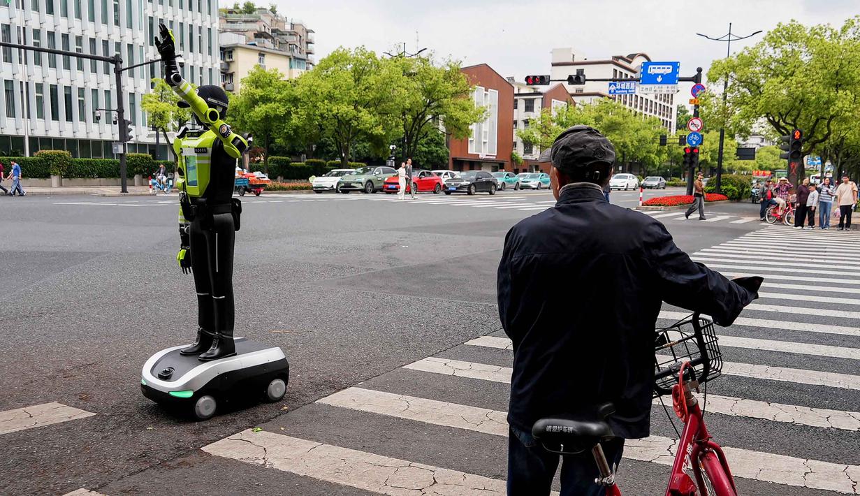 Robot-robot humanoid tersebut diterjunkan untuk membantu kepolisian mengatur arus lalu lintas dan memandu pejalan kaki, khususnya selama libur nasional Hari Buruh yang dimulai sejak 1 Mei 2026. Tampak dalam foto, seorang pesepeda melihat robot polisi humanoid di Hangzhou, provinsi Zhejiang Timur, Tiongkok pada Minggu 3 Mei 2026. (Agatha CANTRILL/AFP)