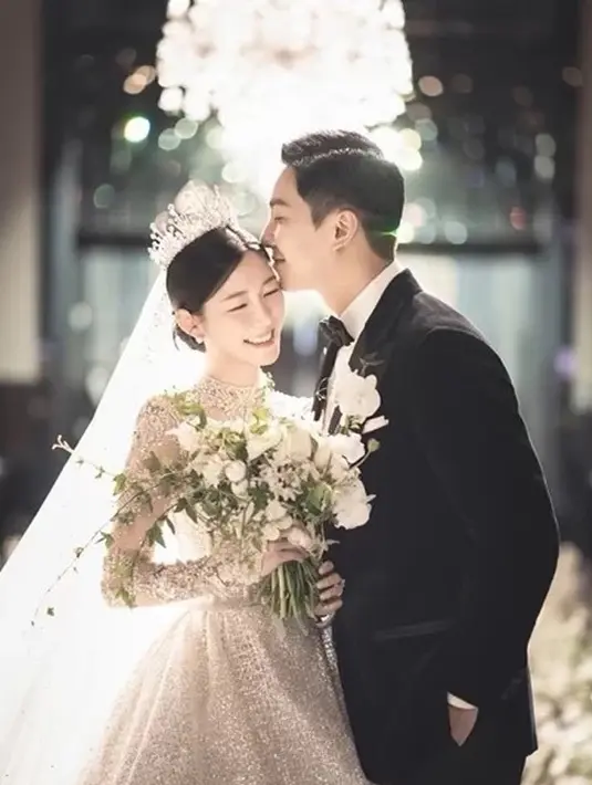 <p>Mengenakan gaun pengantin yang elegan dan tiara mewah, Lee Da In memegang karangan bunga dan tersenyum cerah, sementara suaminya Lee Seung Gi, mengenakan tuksedo di sampingnya, berpose dengan tangan di sakunya dan mencium keningnya. [Foto: IG/byhumanmade]</p>