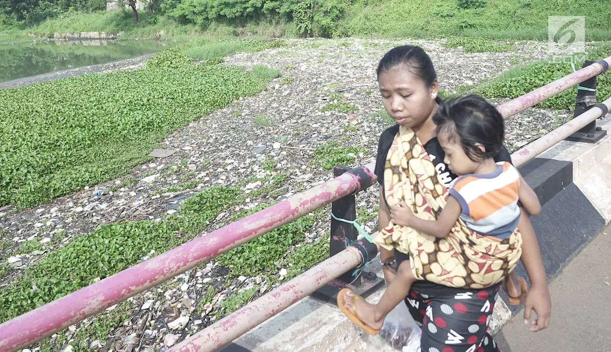 Seorang ibu menggendong anaknya berjalan melintasi Setu Pengarengan yang dipenuhi sampah dan eceng gondok di Depok, Jawa Barat, Selasa (7/5/2019). Kurangnya perawatan menyebabkan Setu Pengarengan kian memprihatinkan. (Liputan6.com/Immanuel Antonius)