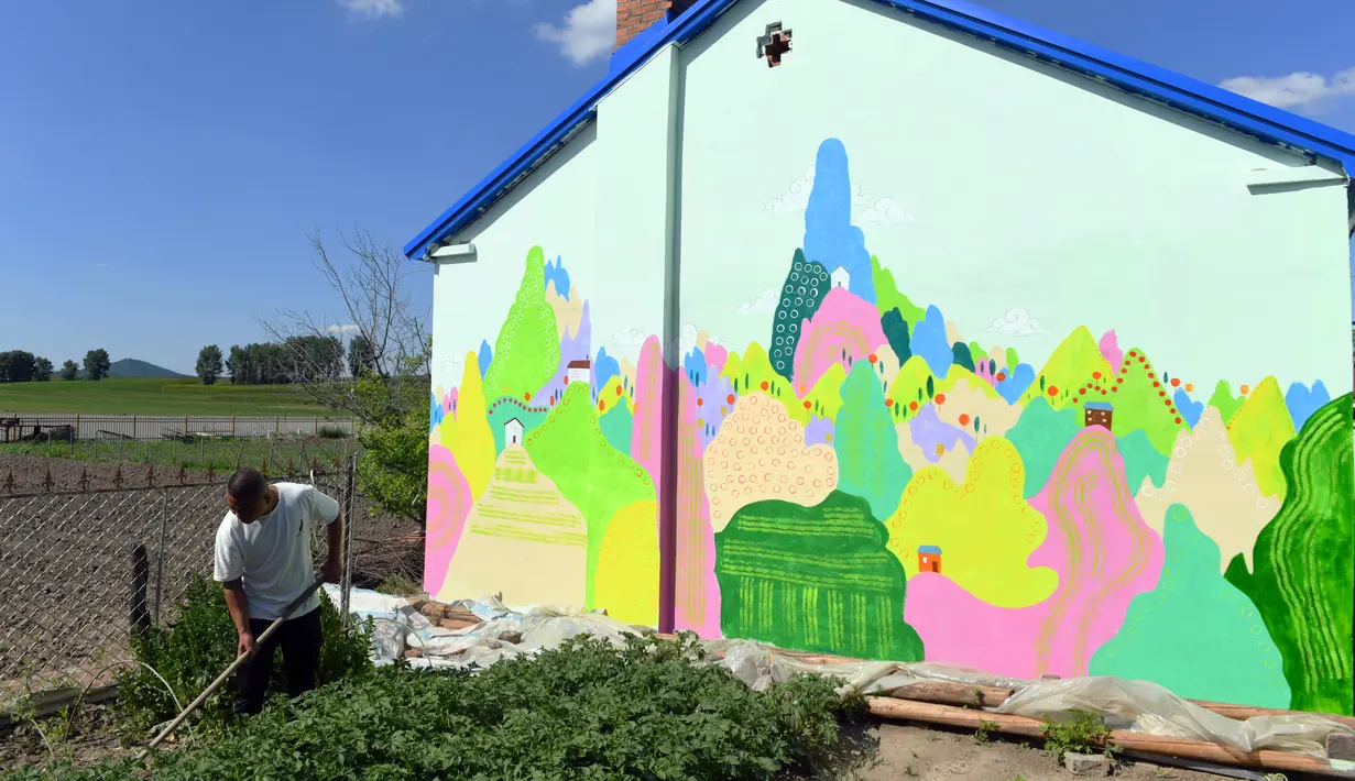 Seorang petani bekerja di samping sebuah rumah yang dihiasi lukisan di Desa Qingshui, Kota Changchun, China timur laut (18/6/2020). Para mahasiswa dan pengajar dari Universitas Seni Jilin menghiasi desa tersebut dengan mural. (Xinhua/Lin Hong)