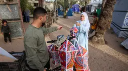 Fanous menjadi simbol suka cita, harapan, dan tradisi yang tetap bertahan di tengah situasi konflik berkepanjangan antara Israel dan Hamas. Tampak dalam foto, Rehan Hazem Sharab, seorang pengungsi Palestina berusia 32 tahun, mengantarkan lampion buatan tangannya, yang dibuat dari kotak-kotak bantuan bekas, sebagai persiapan menyambut bulan suci Ramadan yang dimulai pada 17 Februari, di luar tendanya di lingkungan Al-Mawasi, Khan Younis, di Jalur Gaza selatan pada Sabtu 7 Februari 2026. (Bashar Taleb/AFP)