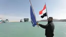 Seorang pria mengibarkan bendera saat kapal kargo Ever Given ditemani kapal tunda melaju di Terusan Suez, Mesir, Senin (29/3/2021). Para ahli percaya efek lanjutan pada pengiriman global bisa memakan waktu berminggu-minggu atau bahkan berbulan-bulan untuk diselesaikan. (Suez Canal Authority via AP)