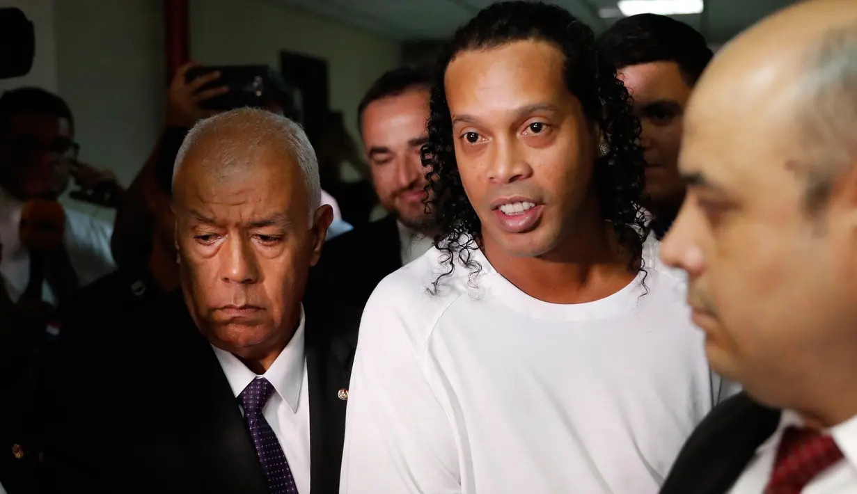Mantan pemain timnas Brasil, Ronaldinho dikawal oleh polisi meninggalkan kantor pengadilan di Asuncion, ibu kota Paraguay, Jumat (6/3/2020). Ronaldinho bersama saudara laki-lakinya berurusan dengan pihak berwenang Paraguay karena menggunakan paspor palsu untuk memasuki negara itu. (AP/Jorge Saenz)