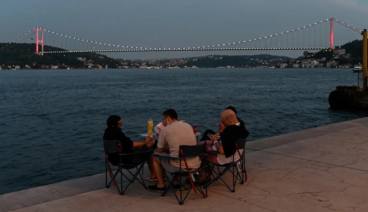 Orang-orang menikmati malam di sebelah pantai Bosphorus ketika jembatan Fatih Sultan Mehmet terlihat di Istanbul (6/7/2020). Jembatan Fatih Sultan Mehmet, yang dikenal dengan sebutan Jembatan Bosporus Kedua, adalah sebuah jembatan di Istanbul, Turki yang melintasi selat Bosporus. (AFP/Ozan Kose)