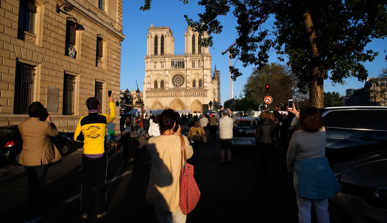 Orang-orang berhenti di depan Katedral Notre-Dame yang kembali membunyikan lonceng utamanya di Paris, Rabu (15/4/2020). Lonceng utama katedral kembali dibunyikan setelah satu tahun mengalami kebakaran yang sampai mengejutkan seluruh warga dunia. (AP/Christophe Ena)