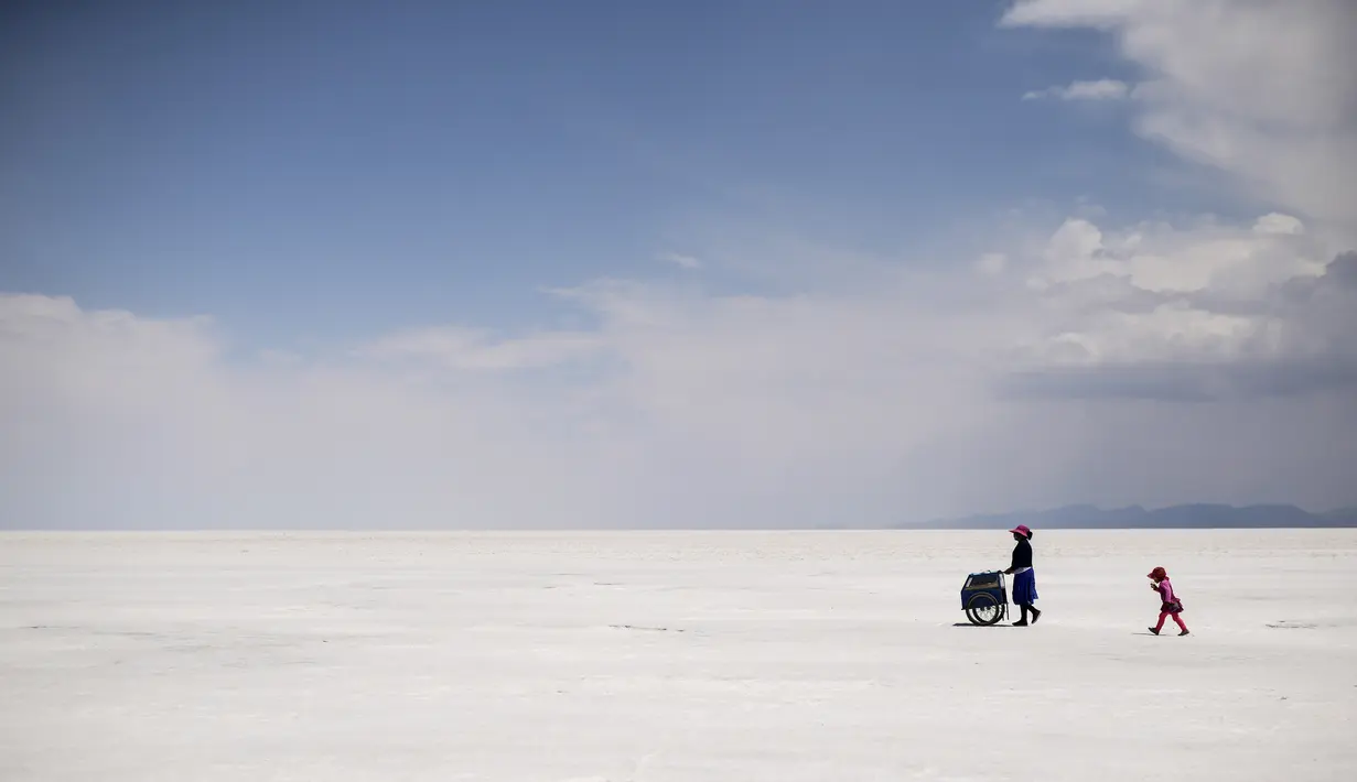 Penjual kerajinan berjalan bersama seorang gadis di Salar de Uyuni, dataran garam terbesar di dunia, di Uyuni, Bolivia, pada 7 November 2020. Dikenal sebagai dataran garam terluas di dunia karena Salar de Uyuni memiliki luas sekitar 4.086 kilometer persegi. (RONALDO SCHEMIDT/AFP)