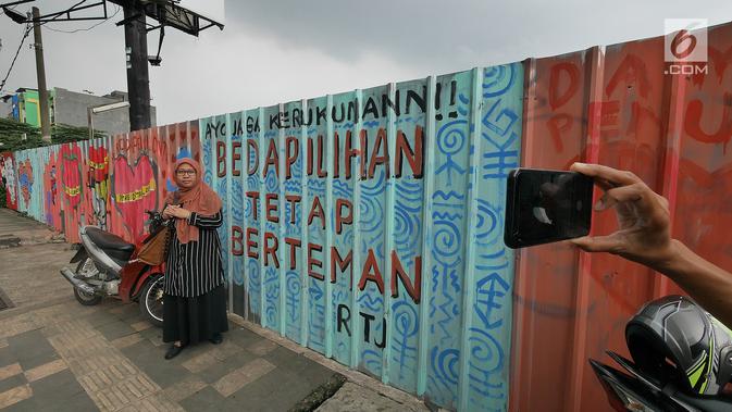 Seorang warga berfoto dengan latar belakang mural di kawasan Margonda, Depok, Sabtu (16/2). Gambar mural memiliki pesan agar masyarakat tetap damai dan berteman meski berbeda dalam memilih calon presiden dalam Pilpres 2019. (Liputan6.com/Herman Zakharia)