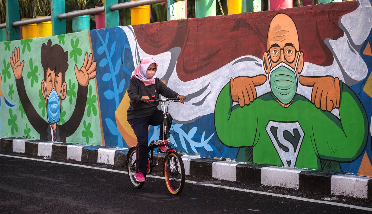 Pesepeda melintas di dekat dinding bermural yang mengajak orang untuk memakai masker di tengah pandemi Covid-19 di Surabaya, Jawa Timur, Minggu (25/10/2020). Dinding itu dipenuhi dengan pesan untuk mematuhi protokol kesehatan di tengah pandemi Covid-19. (Juni Kriswanto/AFP)