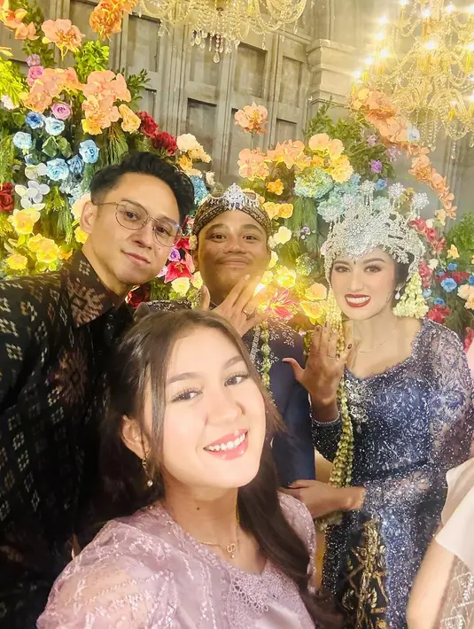 Zoe tampil anggun dalam maxi dress satin warna dusty pink yang dipadukan dengan lace outerwear, memberikan kesan feminin dan elegan. Sementara itu, Antonio memilih mengenakan kemeja batik panjang dengan motif klasik yang dipadukan dengan celana panjang hitam, menciptakan harmoni yang sempurna dalam balutan busana mereka.