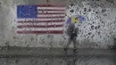 Seorang pejalan kaki membawa payung sambil berjalan melewati lukisan bendera Amerika di San Francisco, Rabu (11/1/2023). California yang dilanda badai berjuang untuk membersihkan dan memperbaiki kerusakan yang meluas. (AP Photo/Jeff Chiu)