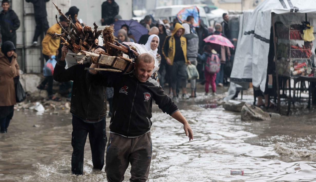 Laporan media internasional menyebutkan cuaca dingin memperberat penderitaan anak-anak dan para lanjut usia di tengah keterbatasan selimut, pakaian musim dingin, serta alat pemanas. Tampak dalam foto, orang-orang membawa kayu sambil berjalan di tengah banjir di kamp darurat yang menampung warga Palestina yang terlantar setelah hujan deras di Kota Gaza pada Selasa 25 November 2025. (Omar AL-QATTAA/AFP)