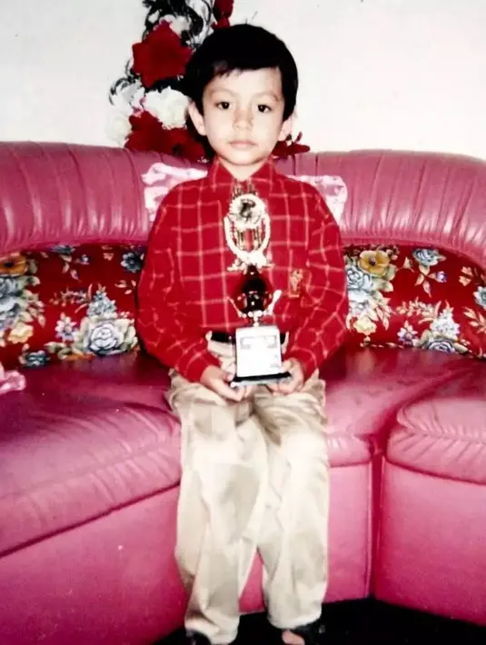 Potret jadul anak kecil dengan memegang piala. Penampilannya tampak polos. Mengenakan baju lengan panjang dan celana panjang duduk di kursi sambil memegang piala. Potret berikut adalah Angga Yunanda saat kecil. [Instagram/@anggayunandareal16-]