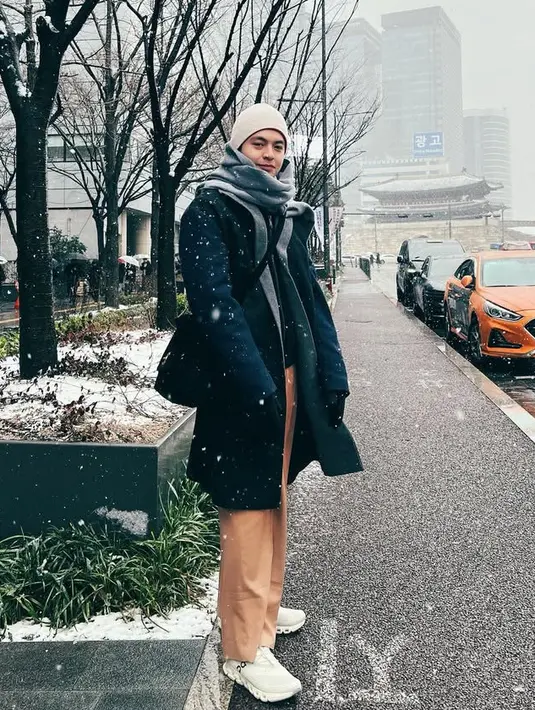 Angga sendiri tampil dengan gaya layering mengenakan inner jaket hitam dengan outer coat warna senada dipadukan celana coklat dan syal abu-abu. Ia pun mengenakan beanie putih dan sling bag hitam. [@angga]