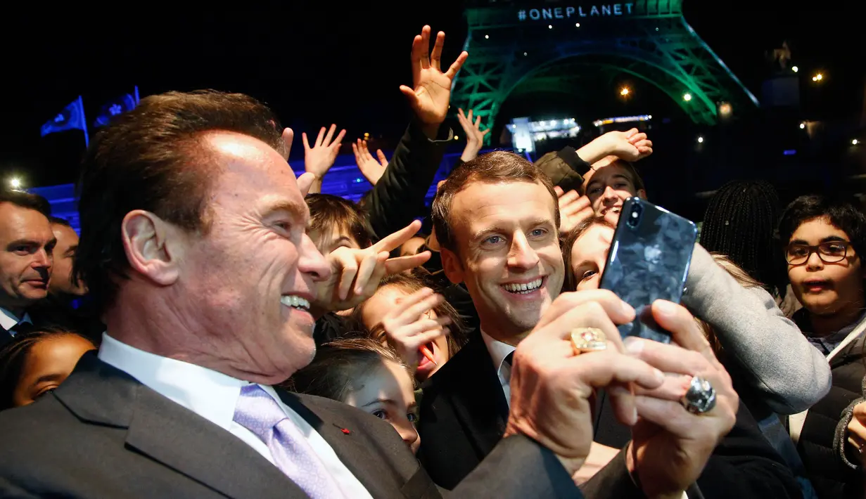 Aktor yang juga mantan Gubernur California, Arnold Schwarzenegger berswafoto bersama Presiden Prancis, Emmanuel Macron dan wisatawan dengan latar belakang Menara Eiffel seusai KTT One Planet Summit di Paris, Selasa (12/12). (AP/Thibault Camus, Pool)