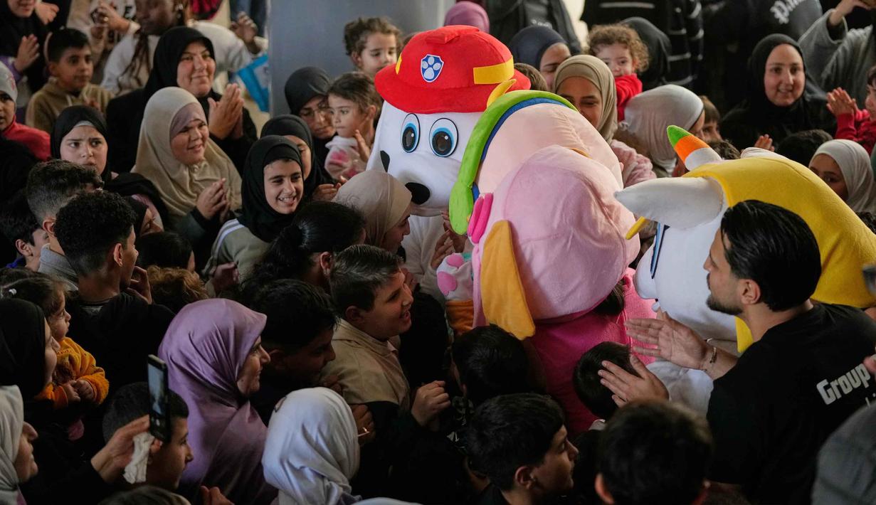 Israel mengklaim serangan tersebut menargetkan infrastruktur dan fasilitas milik Hizbullah guna mencegah serangan roket ke wilayah mereka. Tampak dalam foto, anak-anak berkumpul di sekitar orang-orang yang mengenakan kostum selama acara di mana para sukarelawan menyiapkan bingkisan makanan dan manisan Idulfitri untuk keluarga pengungsi yang merayakan hari raya jauh dari rumah mereka, di sebuah universitas negeri yang diubah menjadi tempat penampungan setelah serangan udara Israel di Beirut, Lebanon, Jumat, 20 Maret 2026. (Foto AP/Hassan Ammar)