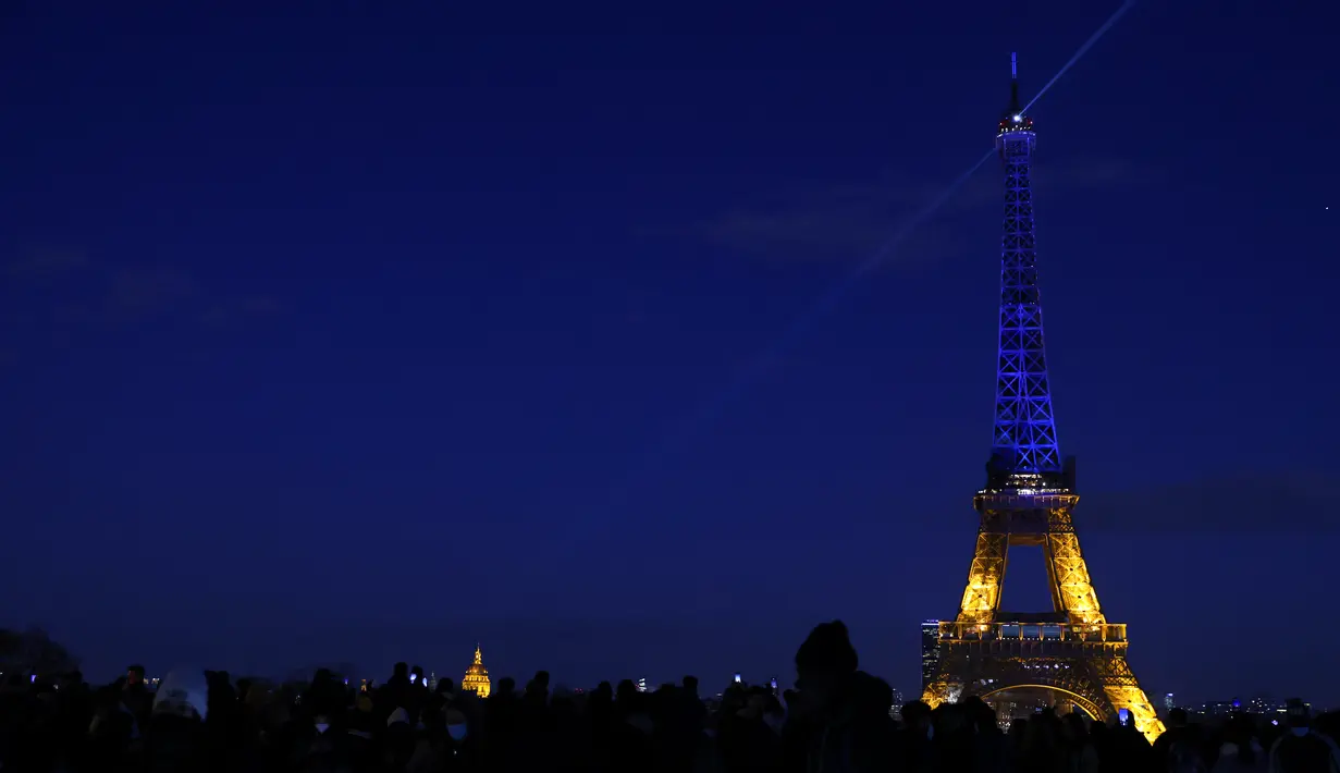 Warga melihat Menara Eiffel menyala dengan warna bendera Ukraina di Paris (25/2/2022). Pasukan Rusia menyerang ibu kota Ukraina pada Jumat (25/2) dengan tembakan dan ledakan dalam invasi ke negara demokratis yang memicu kekhawatiran perang yang lebih luas di Eropa. (AFP/ Thomas Coex)