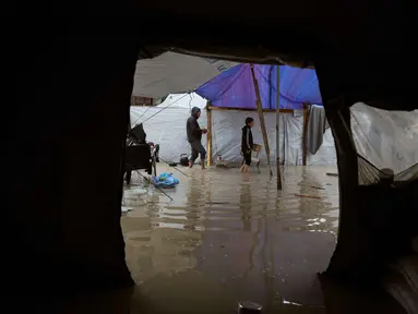 Para pengungsi Palestina berjalan melewati kamp tenda yang basah kuyup akibat hujan lebat di Deir al-Balah, Jalur Gaza tengah, Kamis, 26 Maret 2026. Kondisi kamp pengungsian di Gaza sangat memprihatinkan akibat banjir yang terus berulang selama musim dingin. (AP Photo/Abdel Kareem Hana)