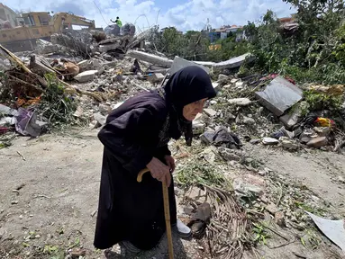 Seorang wanita lanjut usia berjalan melewati petugas pertahanan sipil Lebanon yang sedang mencari korban di antara reruntuhan, sehari setelah serangan Israel di desa Deir Qanun al-Nahr, Lebanon Selatan, pada Rabu 20 Mei 2026. (KAWNAT HAJU/AFP)