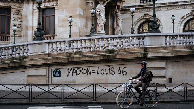 Seorang pengendara sepeda melewati grafiti bertuliskan 'Macron sama dengan Louis 16' di rumah Opera Garnier Paris, Prancis, Minggu (2/12). Hal itu merujuk pada Raja Prancis selama Revolusi Prancis tahun 1789. (AP Photo/Kamil Zihnioglu)
