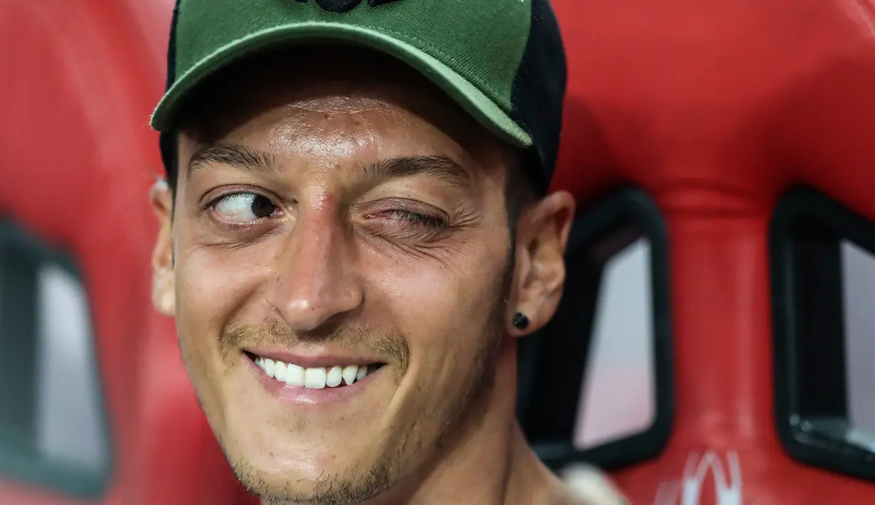 Gelandang Arsenal, Mesut Ozil mengedipkan mata sambil tersenyum ke rekan satu timnya di bangku cadangan selama pertandingan ICC 2018 melawan Atletico Madrid, Singapura (26/7). Arsenal kalah 3-1 setelah bermain 1-1 di waktu normal. (AP Photo/Yong Teck Lim)