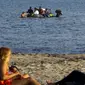 Wisatawan berbaring di pantai melihat para imigran dan pengungsi dari Suriah dan Afrika tiba saat berada diperahu usai melintasi Laut Aegea antara Turki dan Yunani, (8/8/2015). (REUTERS/Yannis Behrakis)