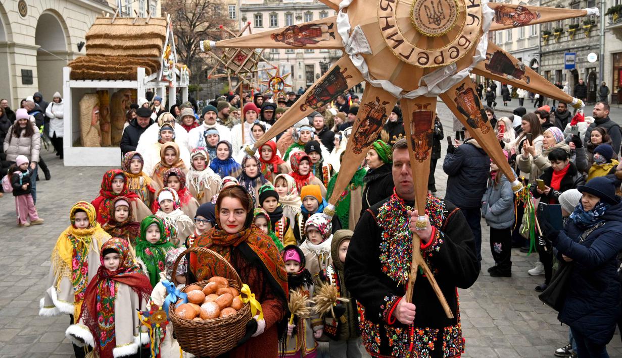 'Didukh' berupa seikat jerami atau gandum yang melambangkan roh leluhur, kemakmuran, dan kesinambungan generasi. Tampak dalam foto, warga Ukraina yang mengenakan pakaian tradisional dan membawa hiasan tradisional Ukraina 'Didukh' menyanyikan lagu-lagu rohani saat mereka mengikuti prosesi Malam Natal di Lviv pada 24 Desember 2025. (YURIY DYACHYSHYN/AFP)