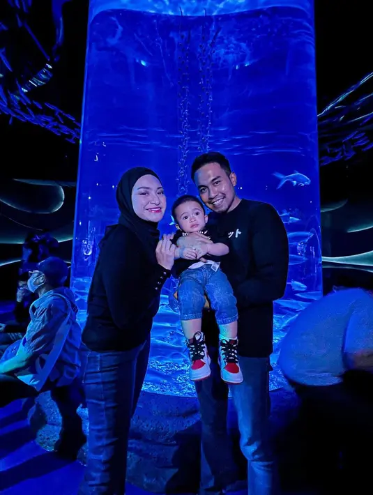 Entah disengaja atau tidak, Nathalie Holscher dan Fariz Utama kompak mengenakan celana jin biru yang dipadukan dengan atasan lengan panjang hitam tanpa kerah. Berfoto dengan latar aquarium vertikal plus tata cahaya lazuardi memberikan efek dramatis remang-remang. Menggendong baby Adzam, keduanya tersenyum lebar menikmati suasana. (Foto: Dok. Instagram @nathalieholscher)