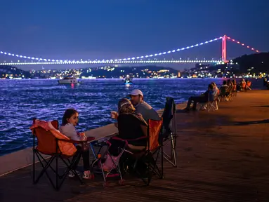 Orang-orang menikmati malam di sebelah pantai Bosphorus ketika jembatan Fatih Sultan Mehmet terlihat di Istanbul (6/7/2020). Jembatan Fatih Sultan Mehmet, yang dikenal dengan sebutan Jembatan Bosporus Kedua, adalah sebuah jembatan di Istanbul, Turki yang melintasi selat Bosporus. (AFP/Ozan Kose)