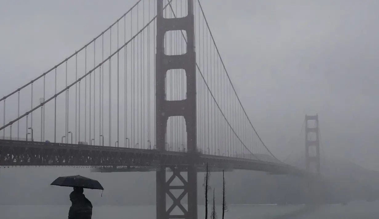 Pejalan kaki berdiri di bawah payung di jalan setapak di depan Jembatan Golden Gate di San Francisco, Rabu (11/1/2023). California yang dilanda badai berjuang untuk membersihkan dan memperbaiki kerusakan yang meluas. (AP Photo/Jeff Chiu)