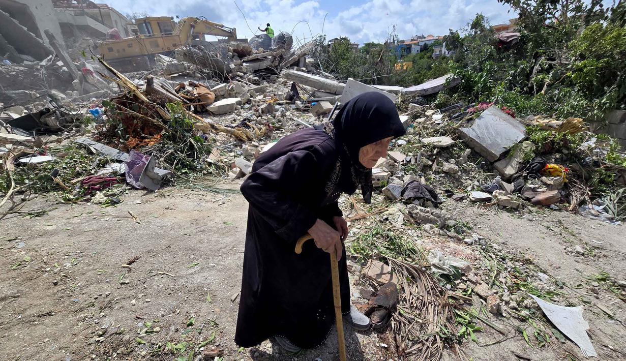 Seorang wanita lanjut usia berjalan melewati petugas pertahanan sipil Lebanon yang sedang mencari korban di antara reruntuhan, sehari setelah serangan Israel di desa Deir Qanun al-Nahr, Lebanon Selatan, pada Rabu 20 Mei 2026. (KAWNAT HAJU/AFP)