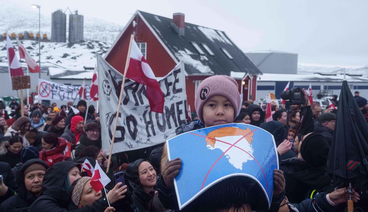Mereka menolak ancaman aneksasi pulau Arktik tersebut. Tampak dalam foto, seorang anak laki-laki memegang peta Greenland yang dicoret dan dihiasi dengan wig yang melambangkan Presiden AS Donald Trump, selama unjuk rasa di depan konsulat AS di Nuuk, Sabtu 17 Januari 2026. (AP Photo/Evgeniy Maloletka)